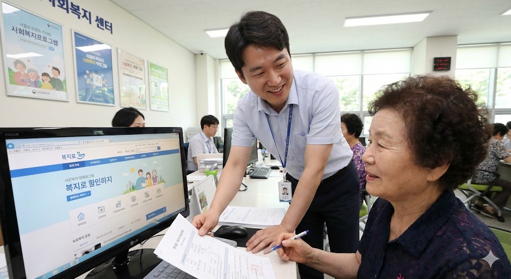 지역사회복지센터에서 한 공무원이 노부인에게 서류와 복지로 웹사이트가 열린 컴퓨터 화면을 보여주며 친절하게 도움을 주고 있습니다. 이는 신청 절차를 상징합니다.