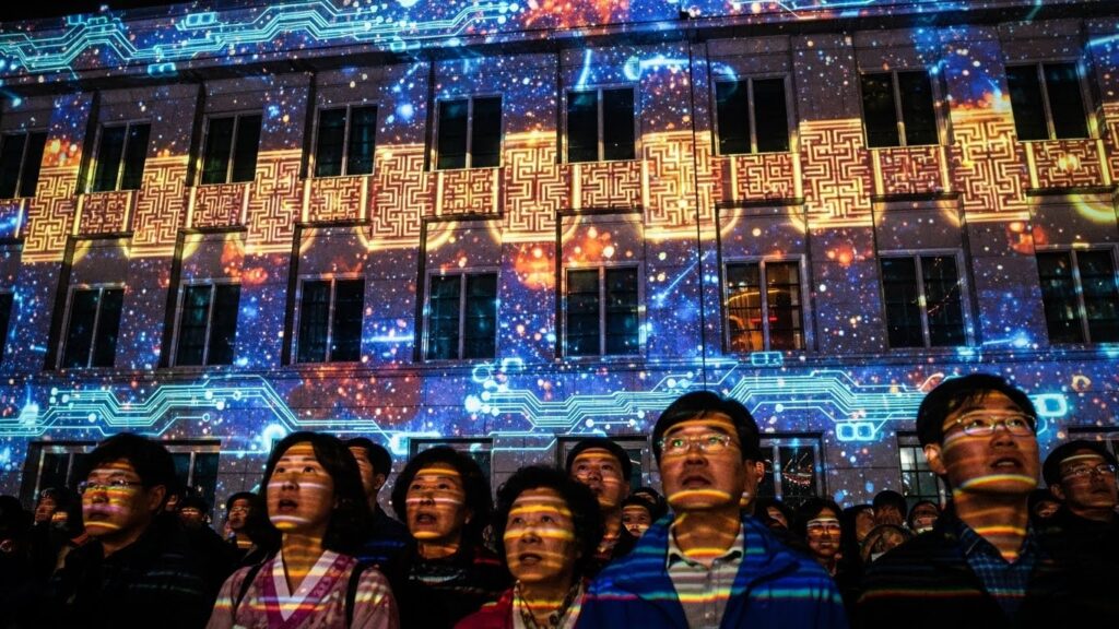 광화문의 한 건물 외벽을 뒤덮은 정교하고 역동적인 프로젝션 맵핑을 가까이에서 촬영했습니다. 전통적인 한국 문양과 미래적인 디지털 아트가 어우러진 화려한 영상에 감탄하는 한국 시민들의 모습이 눈길을 사로잡습니다.